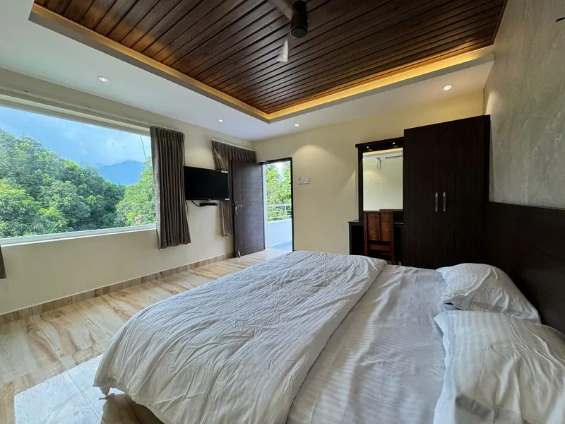 King bed facing a wide picture window with a view of the Western Ghats from a premium AC room at Sai Grand Resorts, Kumbakarai